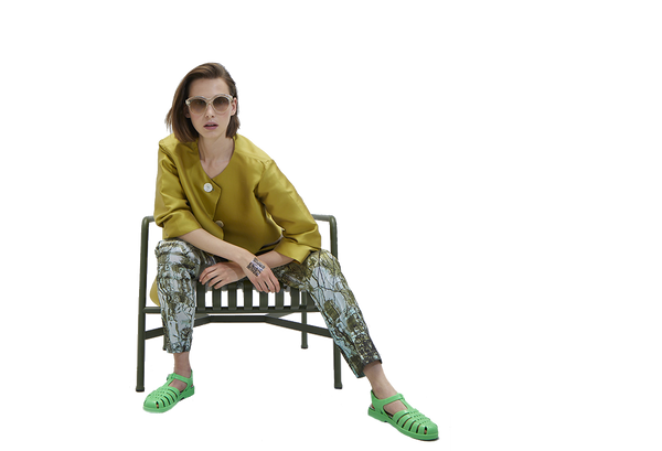 Brünette Frau sitzt auf einem metallischen mattgrünen Gartenstuhl in Seidenhose und -mantel mit giftgrünen Kelly-Sandalen