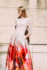 bodenlanger Designerrock mit Artist Print Protea auf cremefarbenen Seidengemisch in Rottönen blondes Model steht an einer Steinwand Alt-Text bearbeiten