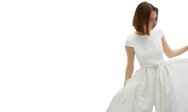 junge Frau mit schulterlangen dunklen Haaren trägt weißes Hosenkleid mit Organza Überrock den sie mit ihren beiden Händen öffnet