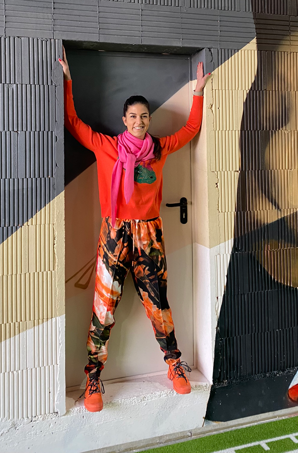 junge Frau steht in Türrahmen von Fitnesscenter trägt eine Jogpant mit dem Muster Azalea und einen passenden Strick Pulli und einen pinken Schal dazu orangene Turnschuhe