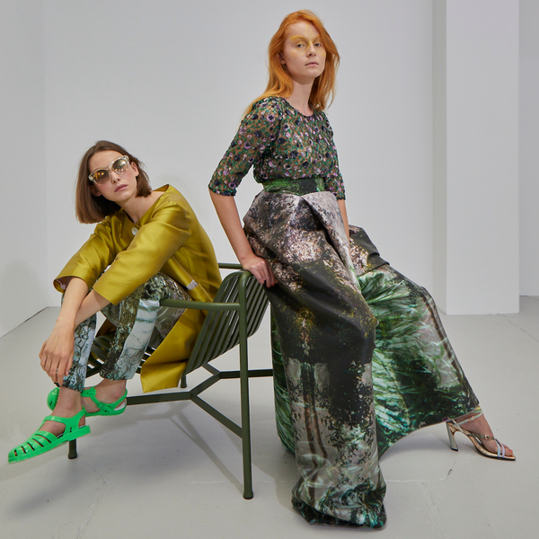 zwei junge Frauen eine rothaarig die anderen braunhaarig sitzend und lehnend an einem grünen metallischen Gartenstuhl und tragen ein halbarm Spitzentop mit Pailletten und bodenlangem Designerrock sowie einem lindgrünen Seidenmantel