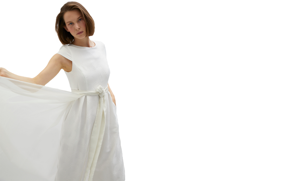 junge Frau mit Kinnlangen brauen Haaren trägt ein weißes Hosenkleid mit Organza Überrock